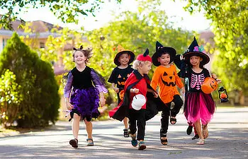 Five children dressed in Halloween costumes, including witches, a skeleton, and a devil, run outdoors on a sunny day holding trick-or-treat buckets.