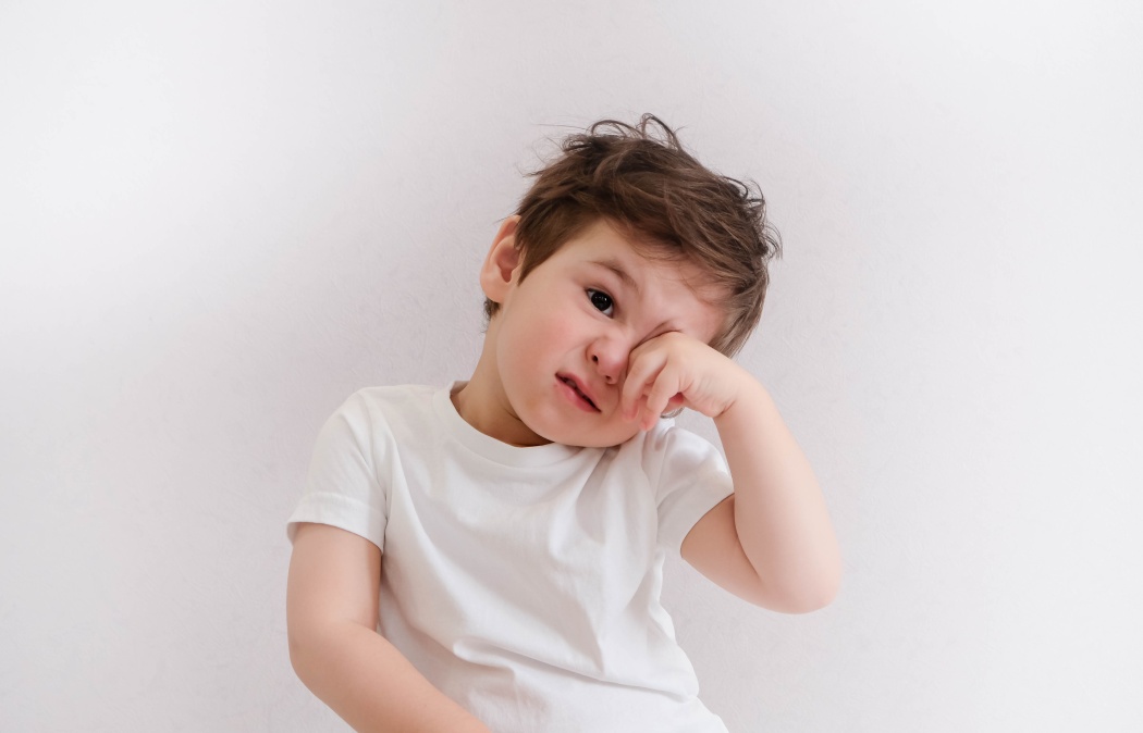 Young child wearing a white t-shirt rubs one eye while looking at the camera against a plain white background.