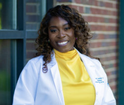 Woman with curly hair wearing a white lab coat and yellow turtleneck stands in front of a brick wall and window, smiling at the camera.