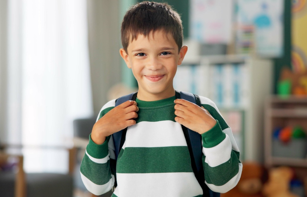 Child, face and laughing in school classroom or education with backpack for kindergarten, learning or joke. Boy, kid and portrait for study development on academy campus or student, humor or funny
