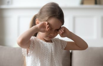 Sleepy stressed tired upset little child crying rubbing eyes feel abused hurt pain, sad lonely worried preschool kid girl in tears miss parents sitting on sofa alone, unhappy children emotion concept