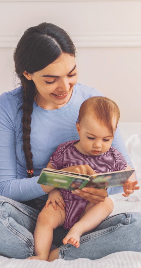Smiling Hispanic Woman Reading Book To Baby While Sitting On Bed At Hom