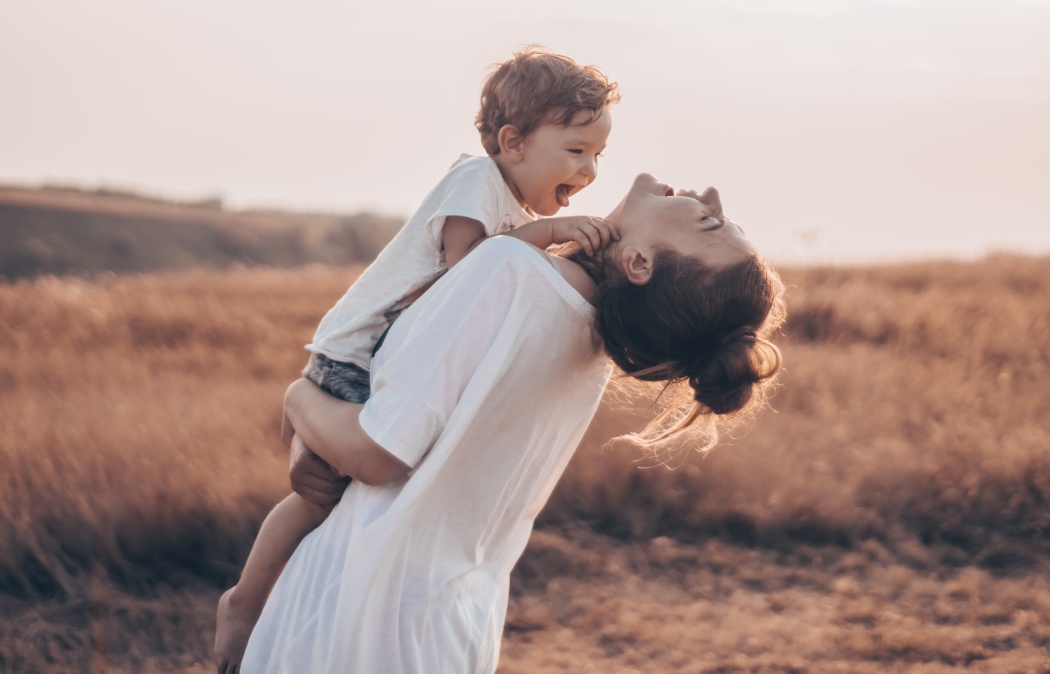 Young woman kisses her little son at sunny in the meadow in sunset. Young mother holding her baby. Mother and little son having good time in the nature. Color toned image. Mothers Day. Family Concept